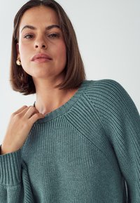 Hellblauer Strickpullover mit gerippter Textur, weitem Ausschnitt und Drop-Shoulder-Design. Goldene Hängohrringe runden den Look ab.