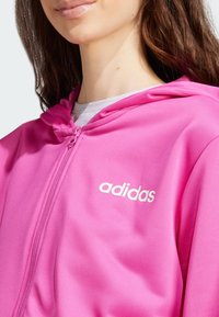Sudadera liviana de color rosa con cremallera, de textura suave y con el logo blanco de "adidas" en la parte izquierda del pecho. Cuenta con capucha y cremallera frontal.