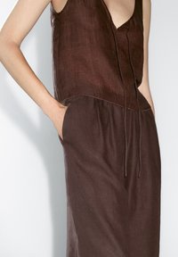 Hnědý lněný top bez rukávů s výstřihem do V a nastavovací šňůrkou; volný střih se bočními kapsami a hladkou, lehkou texturou.