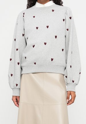 Femme portant un sweat-shirt gris clair avec des motifs de cœurs bordeaux et des manches bouffantes, assorti d'une jupe en cuir beige.