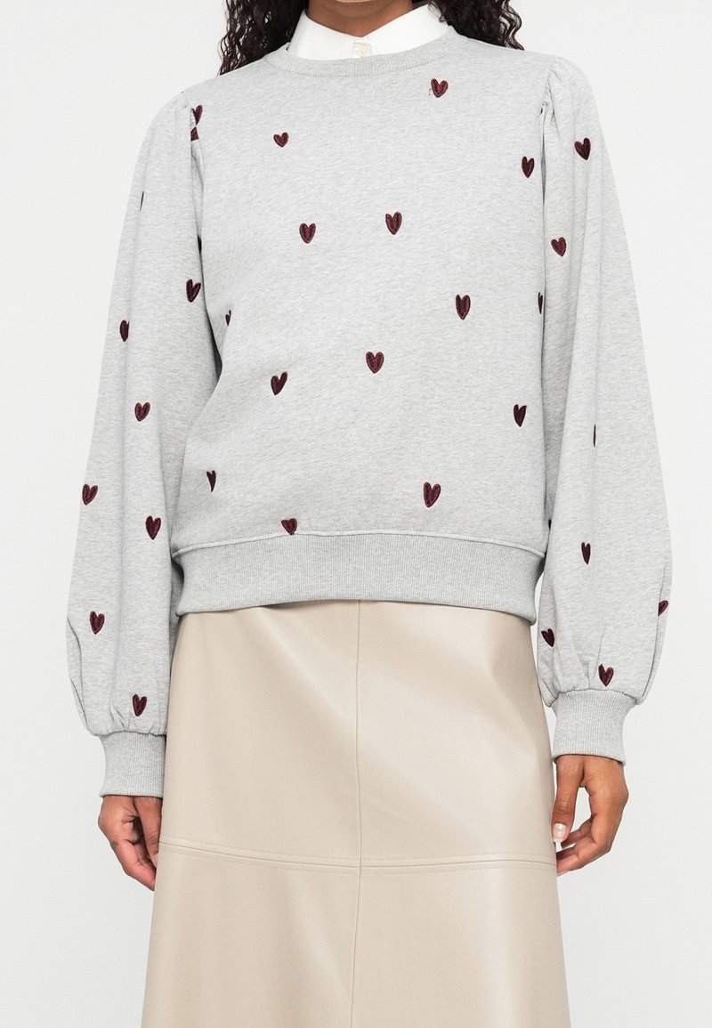 Femme portant un sweat-shirt gris clair avec des motifs de cœurs bordeaux et des manches bouffantes, assorti d'une jupe en cuir beige.