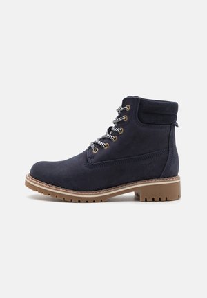 Marineblaue Wildleder-Stiefeletten mit strukturierter Oberfläche, ausgestattet mit Metallösen, grauen Schnürsenkeln und einer braunen Gummisohle mit tiefen Profilen.