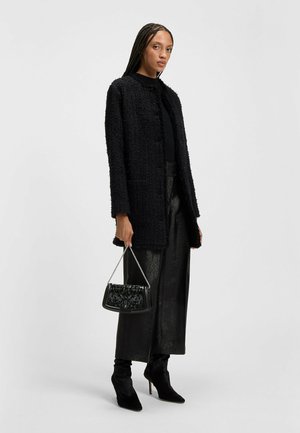 Kvinde med lange fletninger iført en struktureret sort frakke, blanke sorte bukser med bred benvidde, sorte hæle og bærende en sort håndtaske med kædestropp.