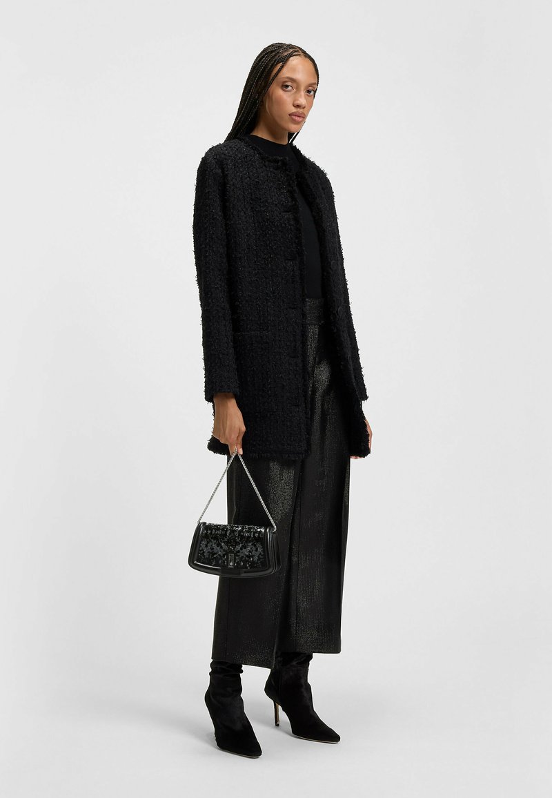 Nainen, jolla on pitkät letit, pukeutunut teksturoituun mustaan takkiin, kiiltäviin mustiin leveälahkeisiin housuihin, mustiin korkokenkiin ja hän pitää kädessään mustaa käsilaukkua, jossa on ketjuolkaimet.