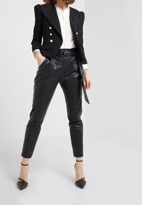 Zwarte op maat gemaakte blazer met gouden knopen, gecombineerd met hooggeplaatste zwarte leren broek en zwarte pumps met spitse neuzen en enkelbandjes.