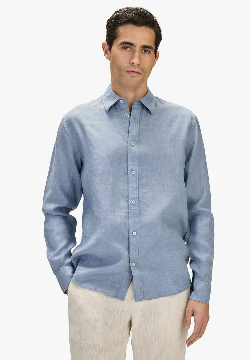 Homme aux cheveux foncés portant une chemise à manches longues boutonnée bleu clair et un pantalon beige, debout avec une main dans la poche devant un fond blanc.