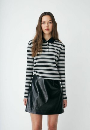Jeune femme aux longs cheveux bruns porte un polo à manches longues rayé noir et gris et une jupe en cuir noire avec des boutons sur le côté.