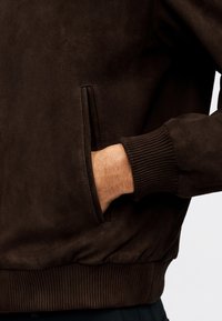 Χέρι σε τσέπη σκούρου καφέ σαμουα φούτερ με ριμπ μανσέτα και μέση, εμφανίζεται από τη μέση μέχρι το μέσο του κορμού.