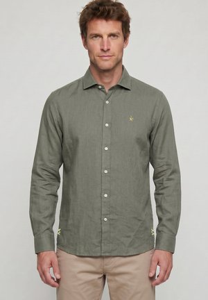 Hombre con camisa de manga larga color verde oliva con botones y pantalones beige, de pie frente a un fondo gris liso.
