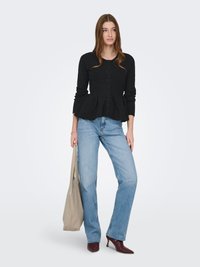 Schwarze Peplum-Strickjacke mit rundem Ausschnitt und Knopfverschluss, kombiniert mit hellblauen Jeans mit geradem Bein und burgunderfarbenen Absatzschuhen, hält eine beige Tragetasche.