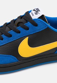 Kék velúr és fekete bőr sneaker sárga Nike logóval, fekete fűzőkkel és párnázott nyakkal. Klasszikus skate design, texturált felületek.