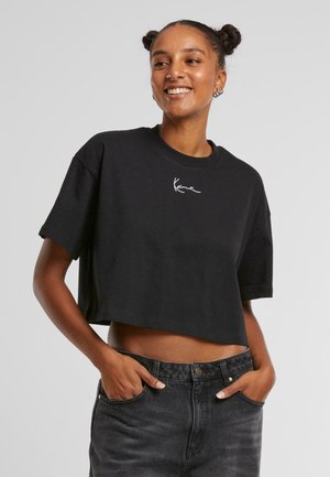 Zwarte cropped t-shirt met korte mouwen en ronde hals. Voorzien van een wit handtekeninglogo op de voorkant. Gecombineerd met donkere spijkerbroeken.