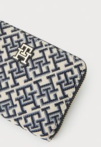 Canvas wallet in cream with navy geometric pattern, featuring a metallic logo and gold zipper. Compact design with textured surface.