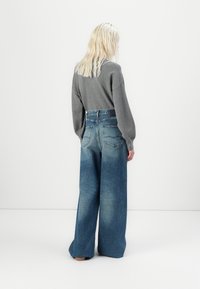 Persoon met lang blond haar, gekleed in een grijze lange mouw top en hoge taille wijdvallende vervaagde blauwe jeans, staand tegen een witte achtergrond.