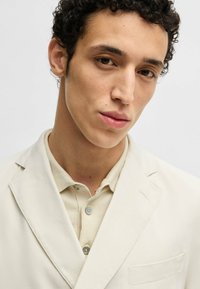 Blazer color beige chiaro con colletto a cono e taschino sul petto, indossato sopra una camicia button-up beige chiaro con una finitura testurizzata.