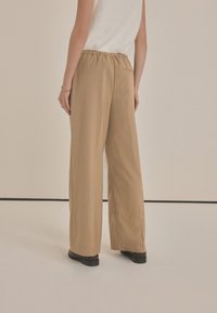 Hochtaillierte weite Hose aus leicht braunem Stoff mit vertikalen Nadelstreifen, einem elastischen Bund und einer Gesäßtasche. Glatte Textur.