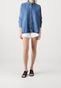 Camicia in denim blu con bottoni e maniche lunghe, abbinata a pantaloni corti bianchi con orlo sfilacciato e mule nere con apertura. Vestibilità semplice e rilassata.