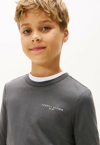 Junger Junge mit kurzen blonden Haaren, der einen dunkelgrauen Tommy Hilfiger Sweater über einem weißen Rundhalsausschnittshirt trägt und leicht lächelt.