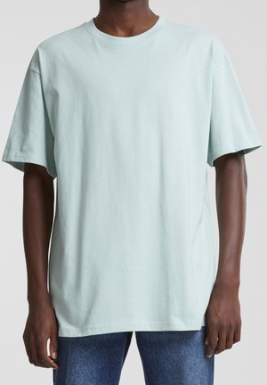 Dunkelhäutige Person trägt ein lockeres, hellblaues Kurzarm-T-Shirt und blaue Jeans und steht vor einem einfarbigen Hintergrund.