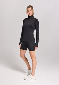 Zwarte langekorte mouwen top met een kwart-rits en aansluitende zwarte shorts, gecombineerd met witte sportschoenen op een lichte achtergrond.