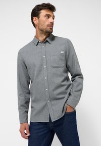 Graues, gewobenes Hemd mit Knopfleiste, einer Brusttasche und einem kleinen Logoetikett, ausgestattet mit einem spitzen Kragen und langen Ärmeln. Kombiniert mit dunkelblauen Jeans.