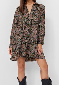 Mujer con un vestido corto de manga larga con un patrón de paisley en verde, naranja y azul, combinado con botas negras hasta la mitad de la pantorrilla.