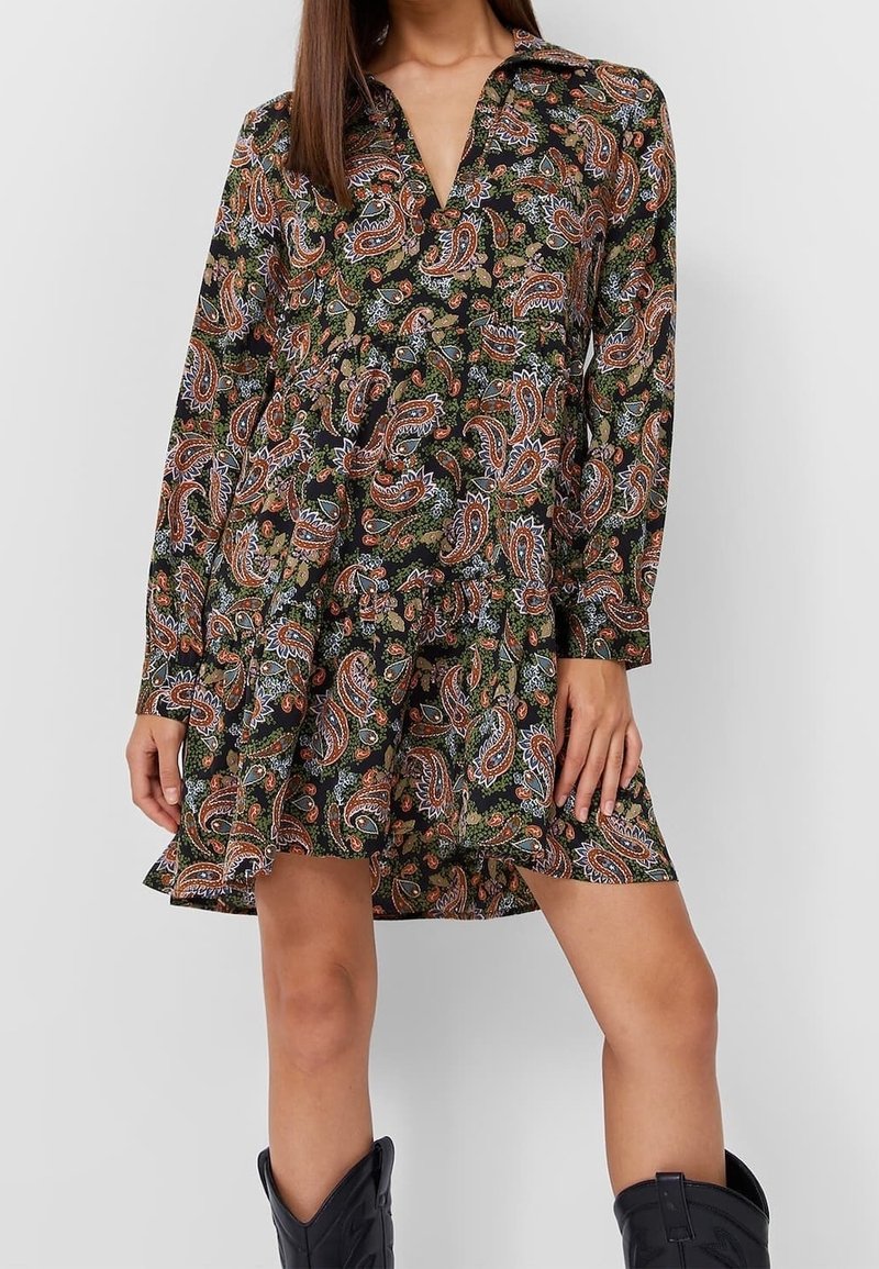 Mujer con un vestido corto de manga larga con un patrón de paisley en verde, naranja y azul, combinado con botas negras hasta la mitad de la pantorrilla.