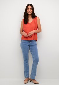Rote ärmellose Bluse mit V-Ausschnitt, kombiniert mit hellblauen Jeans im geraden Schnitt und nude-farbenen High Heels, vor einem neutralen Hintergrund.