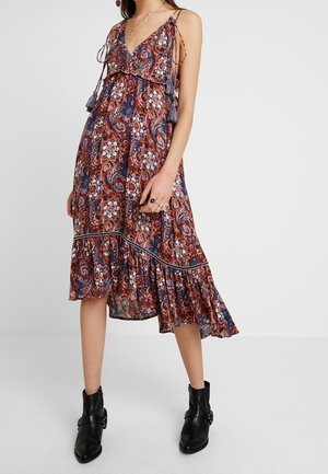 Kobieta w midi sukience bez rękawów, w paisleyowy i kwiatowy wzór, z falbaną u dołu, oraz w czarnych botkach za kostkę.