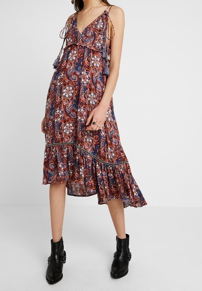 Femme portant une robe midi sans manches à motif cachemire et floral avec un ourlet à volants et des bottines noires.