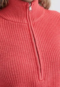 Korallinvärinen neulottu pusero korkealla kauluksella ja vetoketjulla edess ä. Rakenteellisessa kankaassa on pystyribbineulosta ja rento istuvuus.