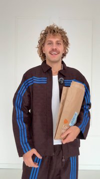 Braune Jacke mit blauen Streifen, strukturiertes Muster, weißes Hemd und braune Papiertüte. Lockere Passform, Kragen und passende Hose sichtbar.