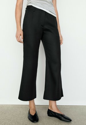 Person trägt schwarze, weit geschnittene, kurze Hosen, schwarze flache Schuhe und ein hellgraues ärmelloses Oberteil vor einem einfarbigen Wandhintergrund.