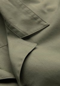 Gros plan sur un tissu vert olive superposé avec des coutures visibles, montrant la texture et les coutures d’un vêtement, probablement une veste ou un manteau.
