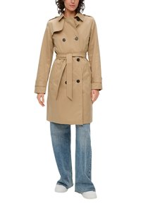 Manteau beige en matière légère avec un devant à double boutonnage, ceinture à la taille et accents de boutons. Le modèle porte un pantalon en denim à jambes larges.