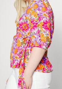 Haut enroulé floral avec des fleurs roses vives, oranges et violettes sur fond blanc ; noué à la taille ; manches courtes bouffantes.