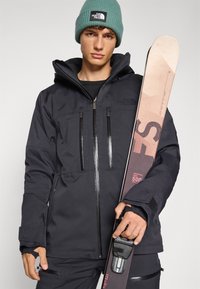 Giacca impermeabile nera con tasche con zip e cappuccio regolabile. Berretto a maglia grigio e sci con un design rosa chiaro e marrone.