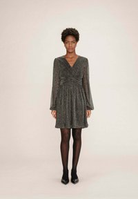 Donna che indossa un vestito scuro grigio lucido, lungo fino al ginocchio e con maniche lunghe, abbinato a collant neri e scarpe nere a punta, in piedi contro uno sfondo neutro.