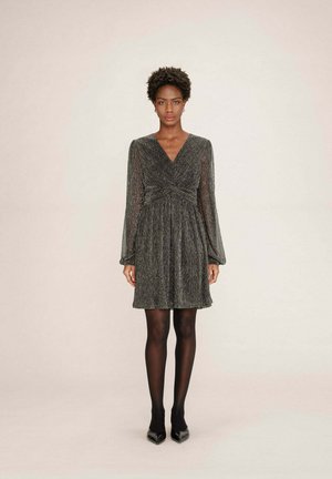 Donna che indossa un vestito scuro grigio lucido, lungo fino al ginocchio e con maniche lunghe, abbinato a collant neri e scarpe nere a punta, in piedi contro uno sfondo neutro.