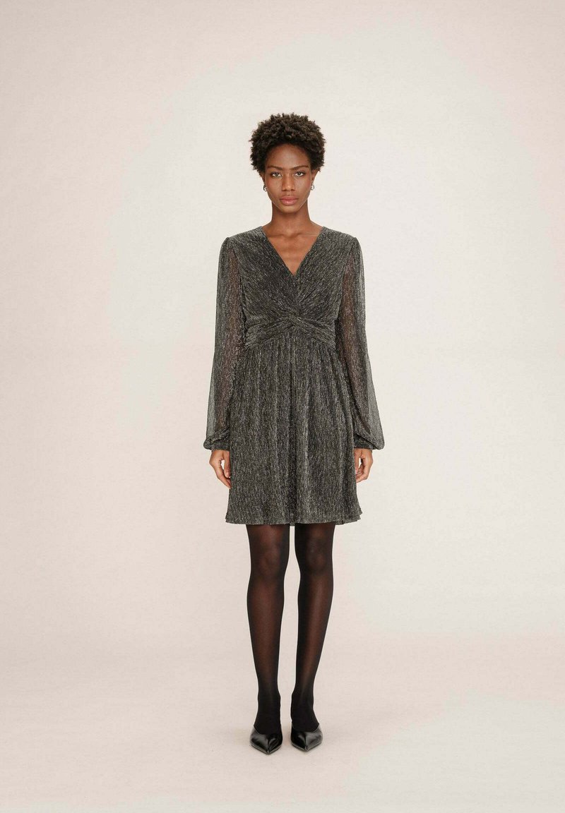 Donna che indossa un vestito scuro grigio lucido, lungo fino al ginocchio e con maniche lunghe, abbinato a collant neri e scarpe nere a punta, in piedi contro uno sfondo neutro.