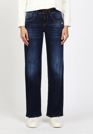 Person trägt dunkelblaue Jeans mit geradem Bein, weiße Sneakers und ein weißes langärmeliges Oberteil und steht vor einem schlichten weißen Hintergrund.