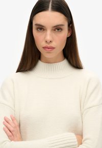 Sweter robiony na drutach w kolorze kremowym, z wysokim rypsowanym kołnierzem i szerokimi rękawami. Miękka tekstura o luźnym kroju. Gładki wygląd tkaniny.