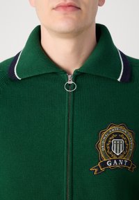 Groene gebreide ritsvest met een groot geborduurd embleem met "GANT" en een marineblauwe en witte gestreepte kraag. Zilveren ritssluiting.