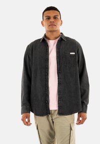 Camisa de mezclilla gris oscuro con botones en el frente, cuello puntiagudo y bolsillo en el pecho izquierdo. Lleva encima una camiseta rosa clara, exhibida sobre un fondo blanco.
