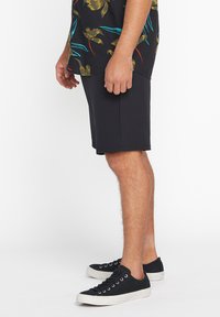 Black shorts made from stretchy material, featuring a mid-thigh cut and side pockets, paired with black sneakers and a floral-patterned shirt.