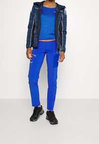 Sininen urheilu takki hupulla ja kontrastivärisin oranssein yksityiskohdin, yhdistettynä sinisiin kapeneviin housuihin, joissa on taskut, sekä mustiin ulkoilukengiin.