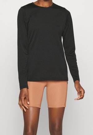 Person iført en sort langærmet top og beige tætsiddende shorts, der står mod en ensfarvet lys baggrund.