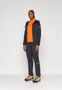 Svart lett jakke med hette, oransje langermet topp, mørk grå bukser, og beige tursko med oransje detaljer; har glidelåslommer.
