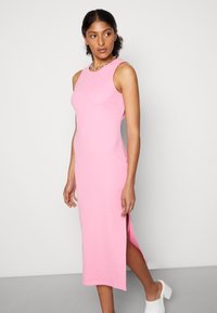Vestido midi sem mangas, cor-de-rosa, com pregas e fenda lateral; o design ajustado realça a silhueta. Modelo usa mules brancas de salto e um colar de corrente.