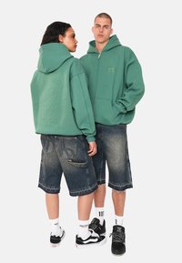 Grüne Zip-Hoodies mit geprägten Logos, kombiniert mit blauen Jeansshorts mit ausgefranstem Saum und sichtbaren Taschen. Schwarze Turnschuhe runden den Look ab.